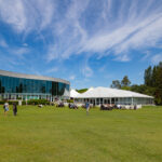 People sitting and walking on a large grassy field in front of a curved modern building and a white tent under a blue sky with wispy clouds.