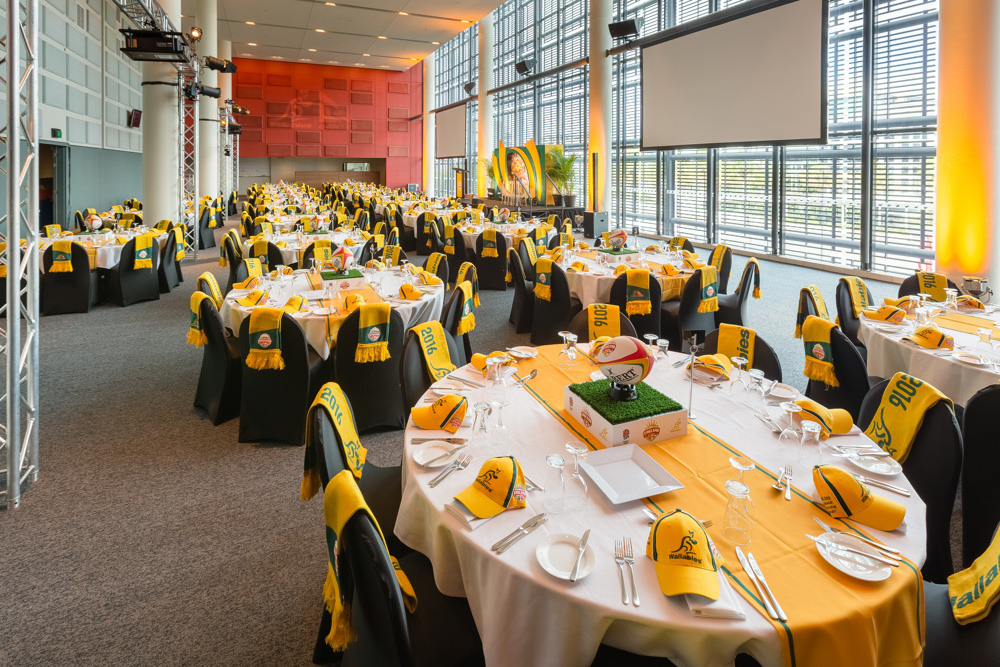 Spacious event hall set up for a sports-themed banquet with yellow and black decorations, tables arranged with glasses, plates, and rugby-themed centerpieces.