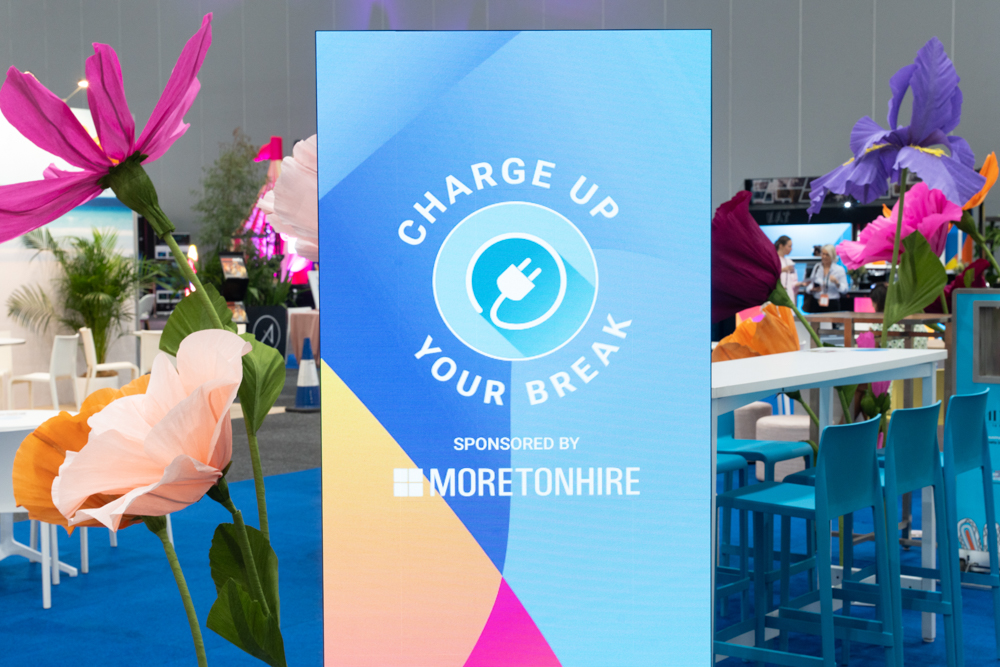 Sign displaying "Charge Up Your Break," sponsored by Moreton Hire, amidst colorful oversized flowers and event seating.