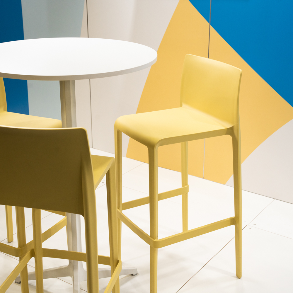 A modern, yellow bar stool is set next to a round white table against a geometric blue and yellow wall background.