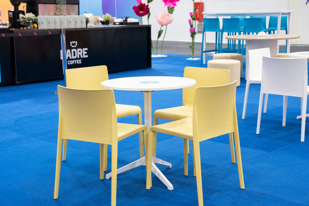A small round table with four yellow chairs is on a blue carpet. In the background, there's a black coffee counter and more tables and chairs.
