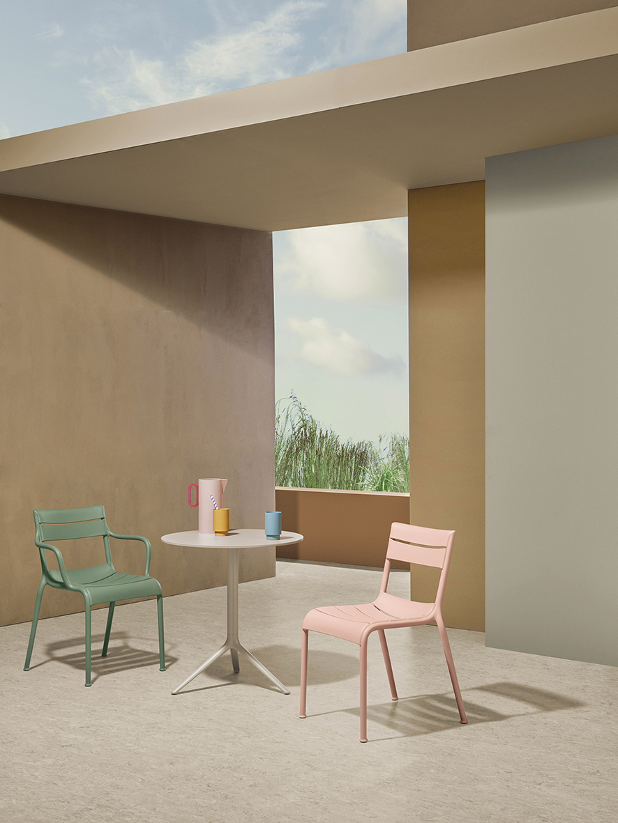 Two modern chairs, one green and one pink, are placed around a small round table with colorful mugs on it, set in a minimalist outdoor setting with beige walls and a view of greenery.