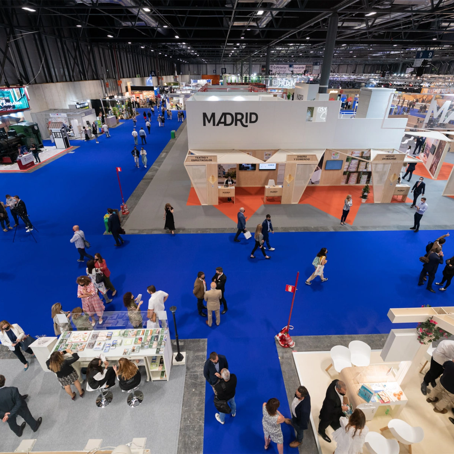 Aerial view of a trade show with people walking between booths. A large booth with the word "Madrid" is prominently displayed in the center on blue carpeted flooring.